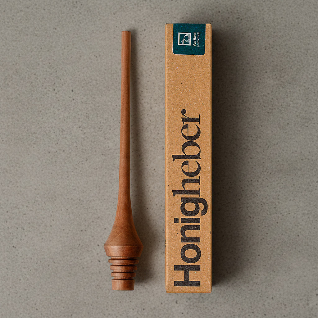 Ein Honiglöffel aus Holz, der aufrecht liegt. Der Löffel hat einen langen, schlanken Griff und am unteren Ende befinden sich mehrere Einkerbungen für das Aufnehmen von Honig. Die Oberfläche des Löffels ist glatt und poliert mit einem Bienenwachsfinish. Handgemachter Honigheber aus Elsbeere. Danben liegt die dazugehörige Verpackung von der tischlerei Mimikry