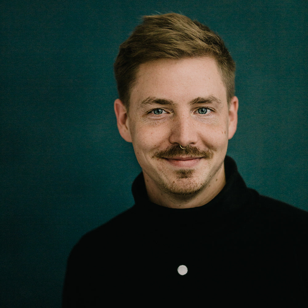 Porträt von Niklas Deinzer, lächelnd, mit kurzem, hellbraunem Haar und Oberlippenbart vor einem dunklen Hintergrund im mimikry, dem designbüro aus Leipzig