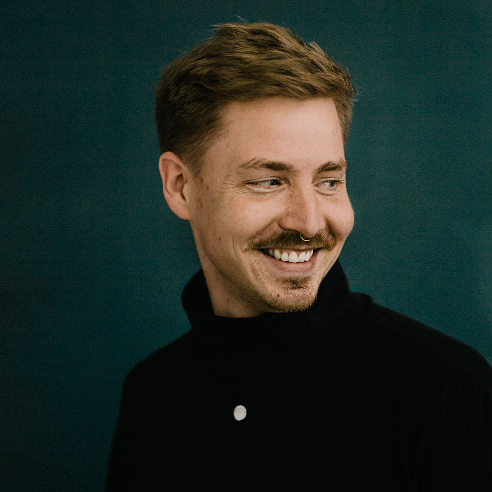 Porträt von Niklas Deinzer, lächelnd, mit kurzem, hellbraunem Haar und Oberlippenbart vor einem dunklen Hintergrund im mimikry, dem designbüro aus Leipzig. Er schaut nach unten links und strahlt.