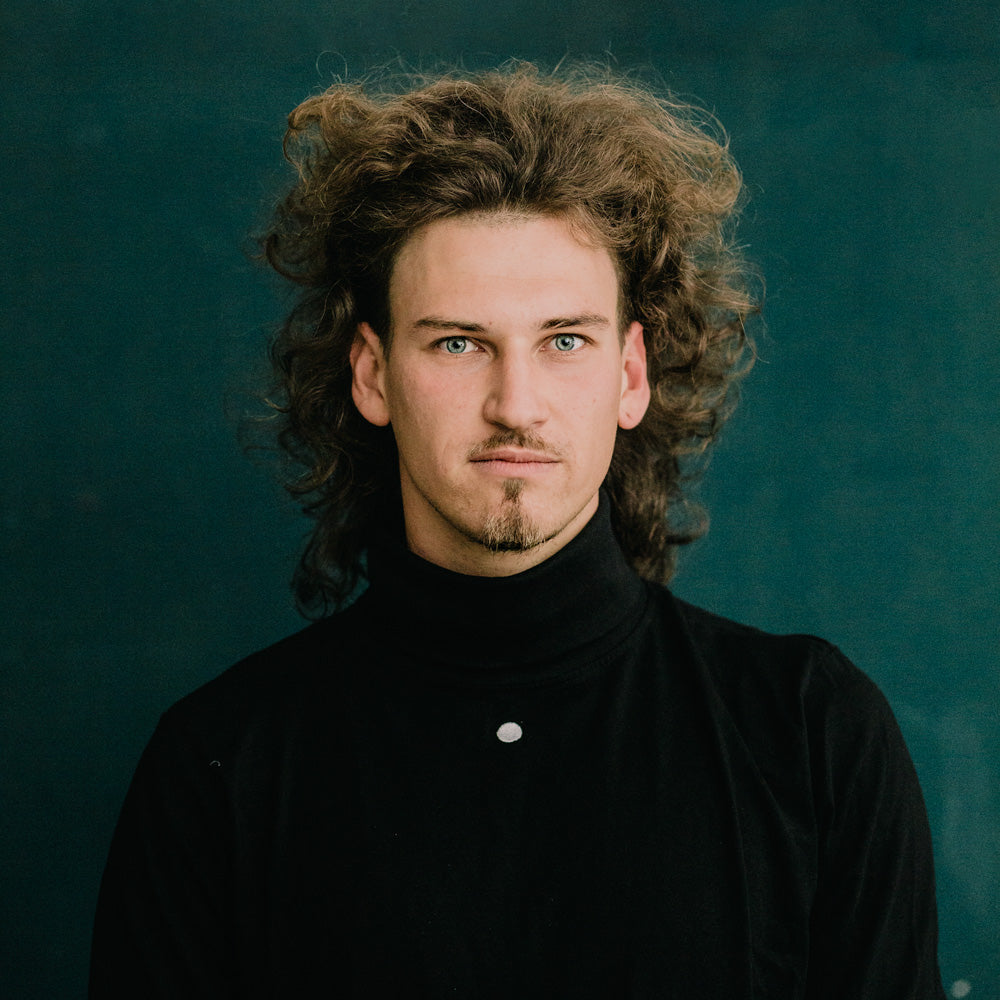 Porträt von Paul Rothfuß, mit lockigem, langem Haar und dunklem Rollkragenpullover vor einem dunklen Hintergrund im mimikry, dem Designbüro in Leipzig