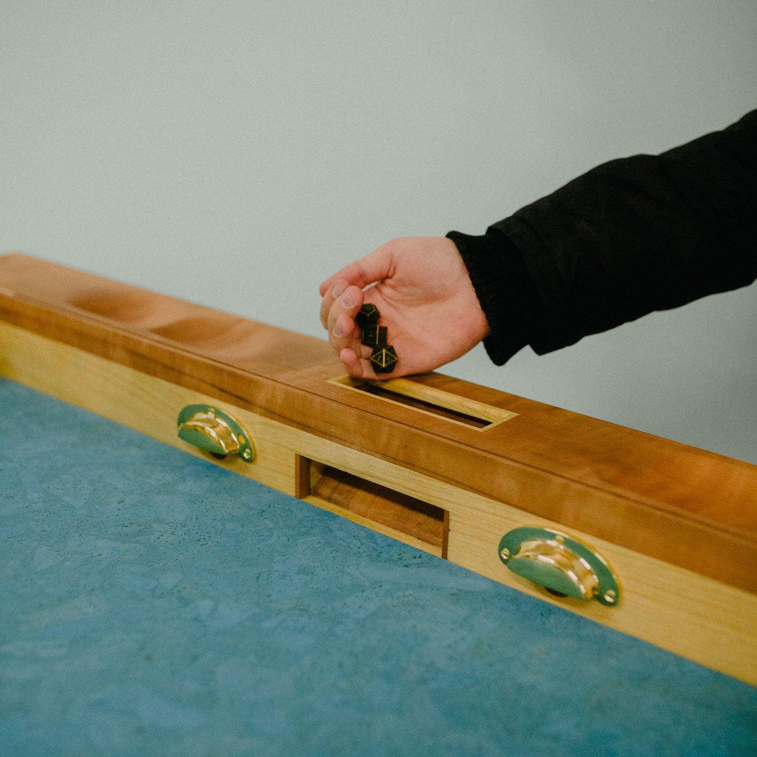 Detailaufnahme einer Hand, die Würfel durch den Würfelturm des Spieletisches Corbin wirft. Der Spieletisch besteht aus Elsbeere und Kirsche. Messingdetails werten das Designmöbel weiter auf.