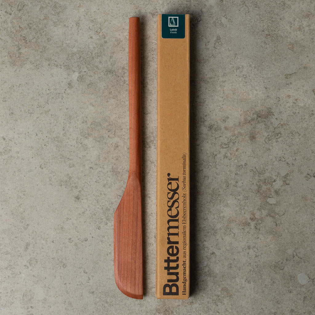 Buttermesser aus Holz, aus Elsbeerenholz gefertigt von Leipziger Tischlerei mimikry, neben dessen Verpackung für manufactum