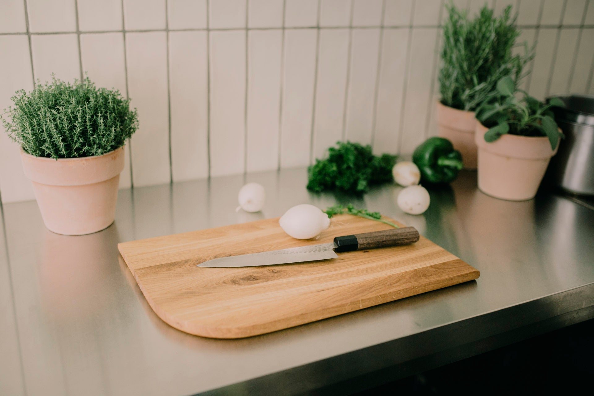 Ein großes Schneidebrett aus Eiche liegt auf einer Edelstahlküchenfläche, umringt von Gemüse und Frischen Kräutern