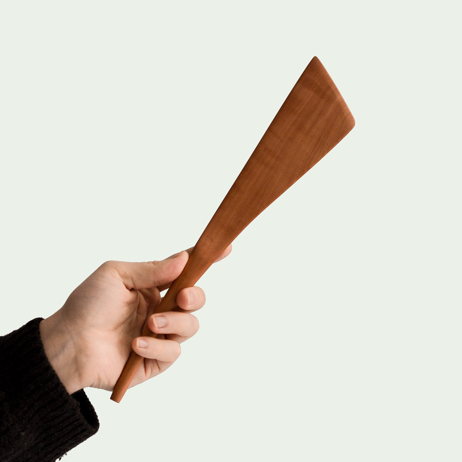Hand hält handgefertigten Pfannenwender aus Elsbeere mit breitem, flachem Design und glatter Oberfläche, nachhaltig produziert, diagonale Ansicht.
