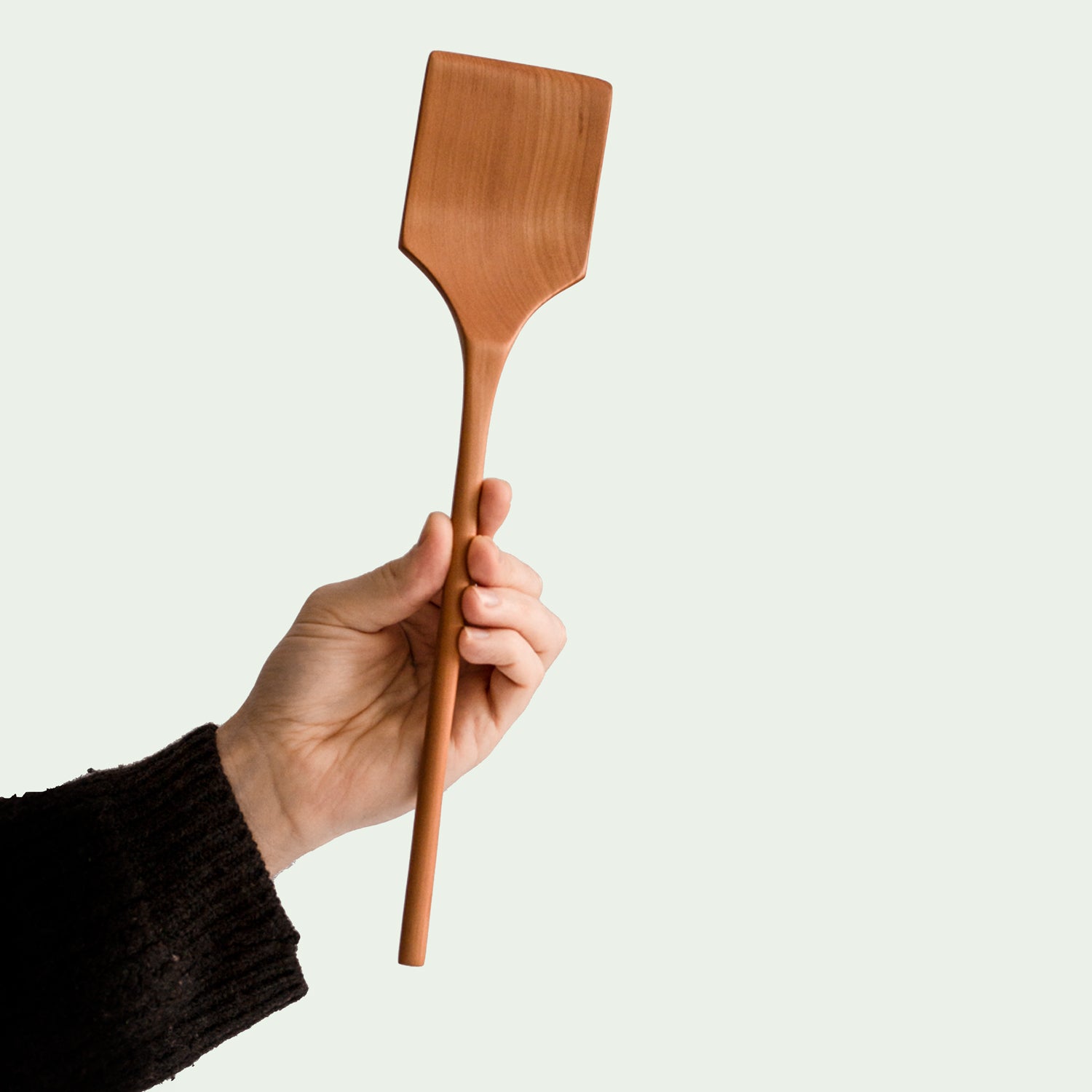 Hand hält handgefertigten Pfannenwender aus Elsbeere mit rechteckiger, leicht gebogener Schaufel und ergonomischem Griff, nachhaltig produziert, diagonale Ansicht.