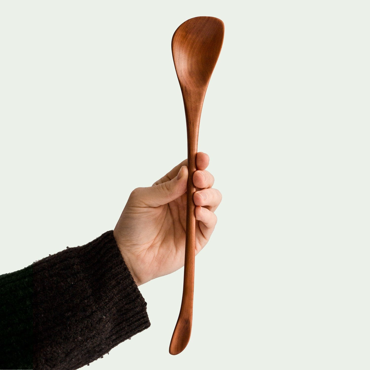 and hält handgefertigten asymmetrischen langen Löffel aus Elsbeere mit elegant geschwungenem Design und glatter Oberfläche, nachhaltig produziert.