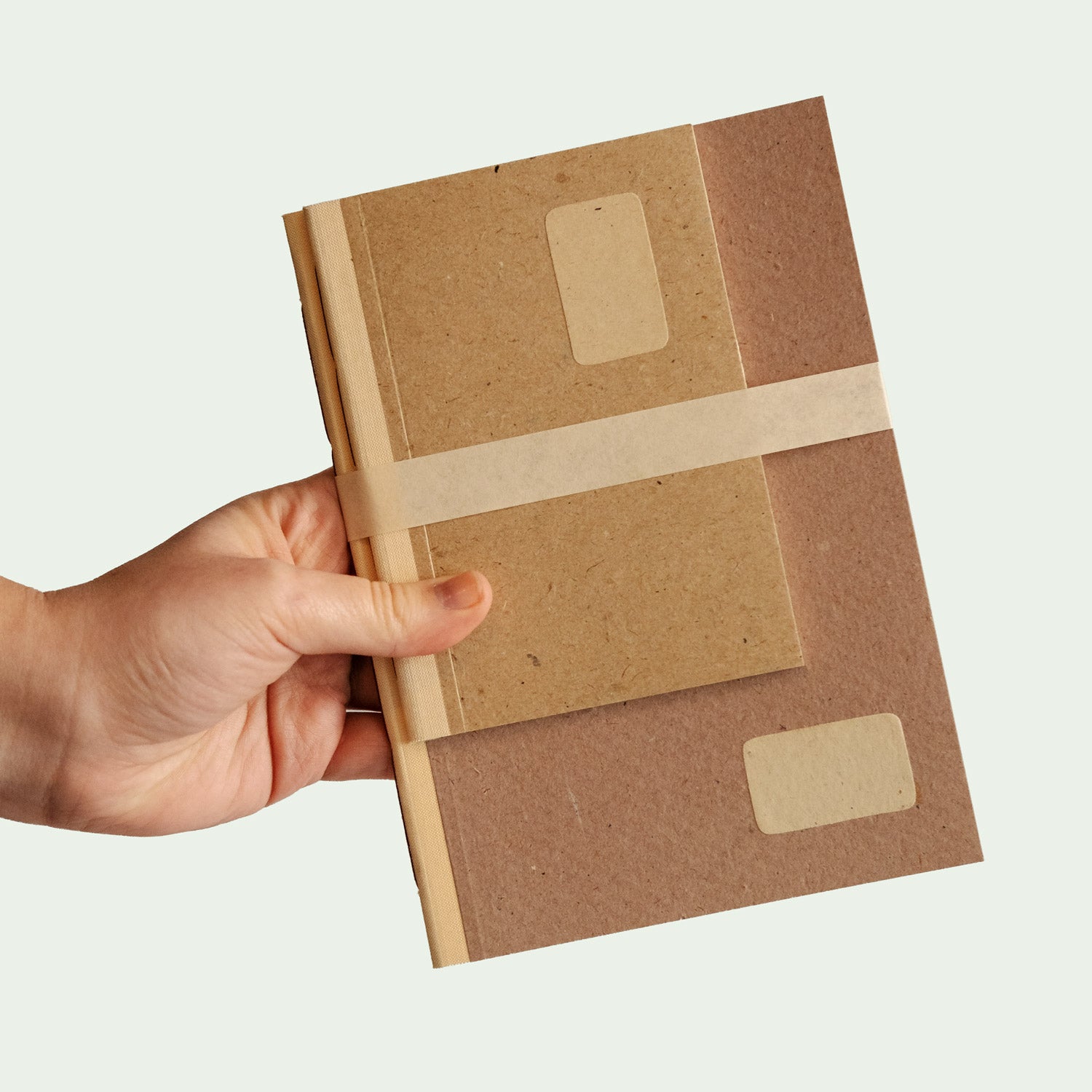 Eine Hand hält ein zweiteiliges Notizbuch-Set. Das größere Buch ist braun, während das kleinere hellbraun ist. Beide Bücher sind mit einem hellen Band zusammengebunden.