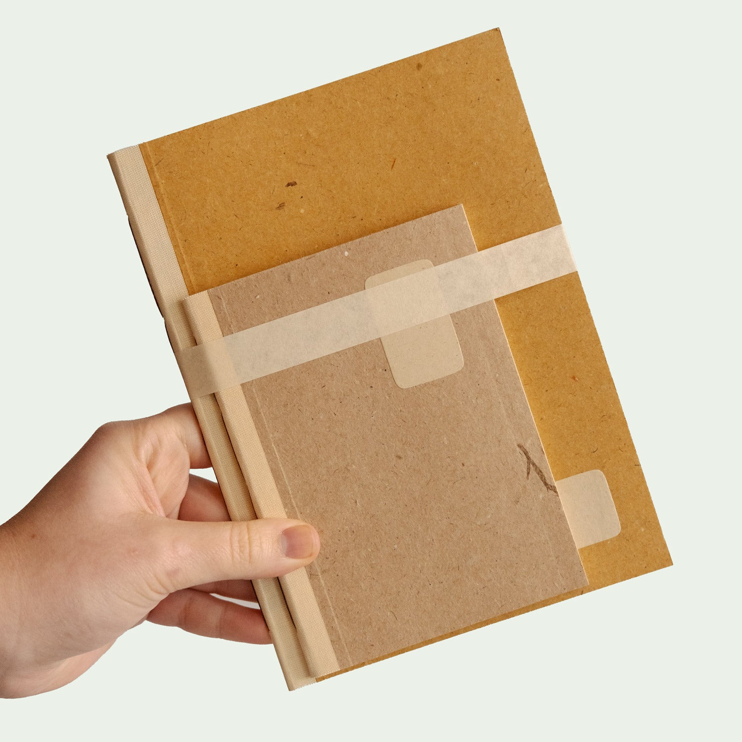 Eine Hand hält ein zweiteiliges Notizbuch-Set. Das größere Buch ist Gelb, während das kleinere hellbraun ist. Beide Bücher sind mit einem hellen Band zusammengebunden.