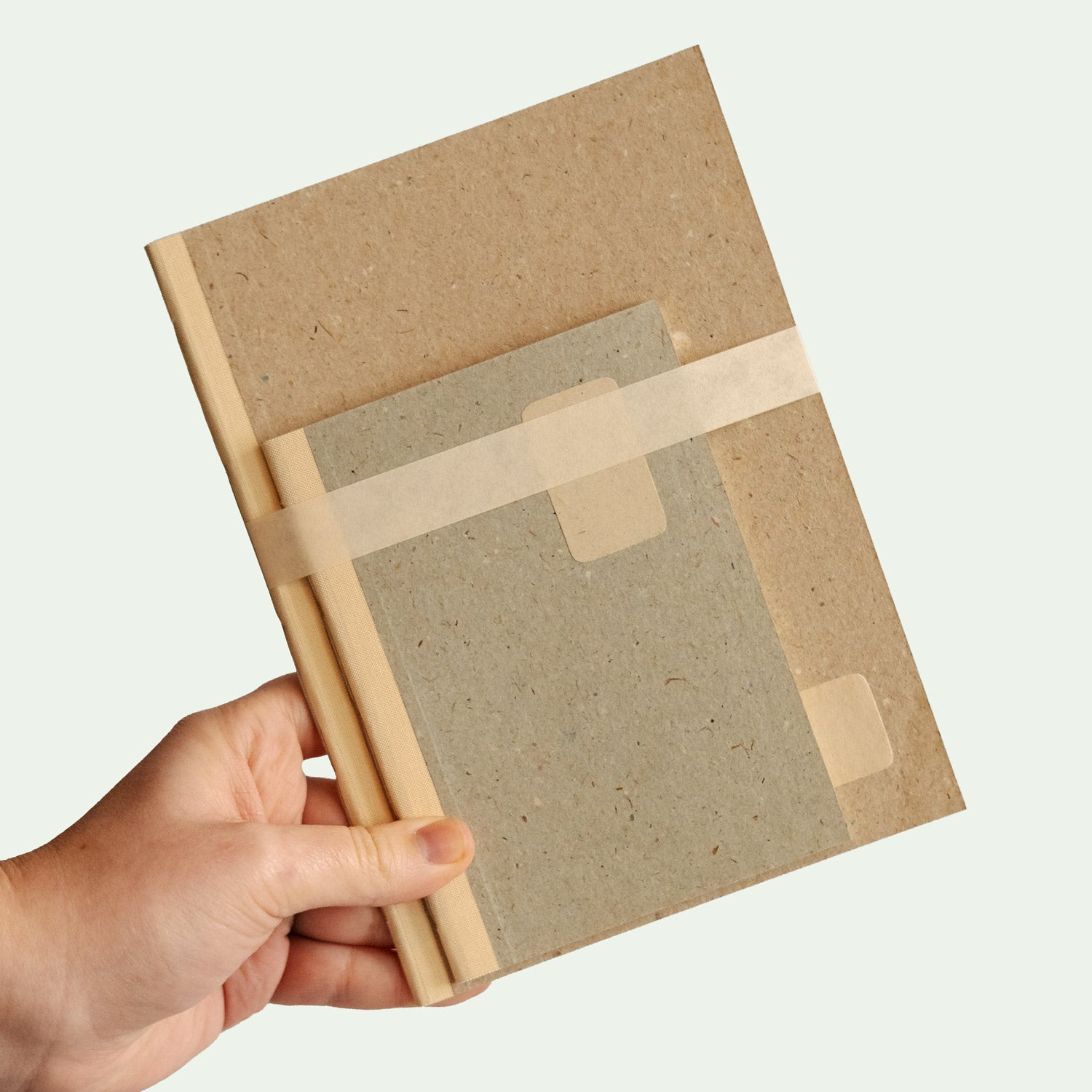 Eine Hand hält ein zweiteiliges Notizbuch-Set. Das größere Buch ist beige, während das kleinere grau ist. Beide Bücher sind mit einem hellen Band zusammengebunden.