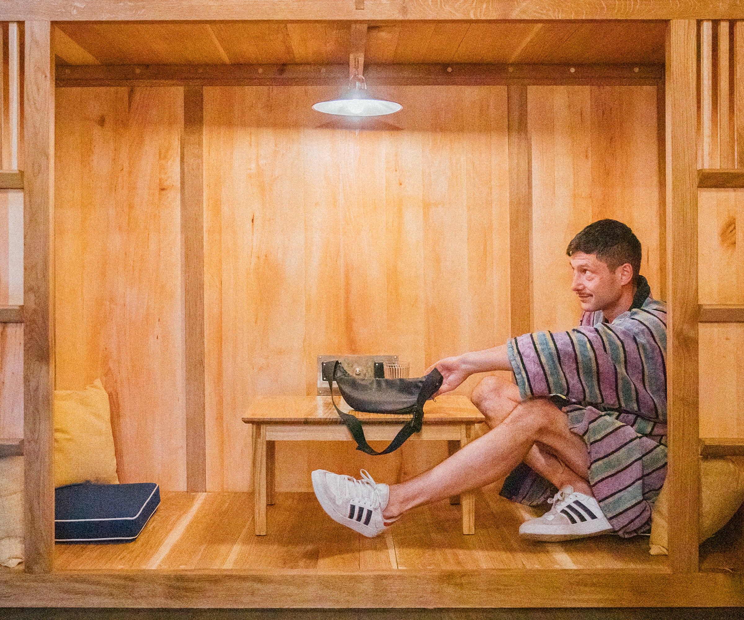 Eine Person sitzt in einer hölzernen Nische, trägt einen bunten Bademantel und weiße Adidas-Sneaker. Die Nische ist mit einem Tisch, einer Lampe und Steckdosen ausgestattet. Die Sitznische wurde von mimikry, einem Designbüro aus Leipzig entworfen und gebaut.