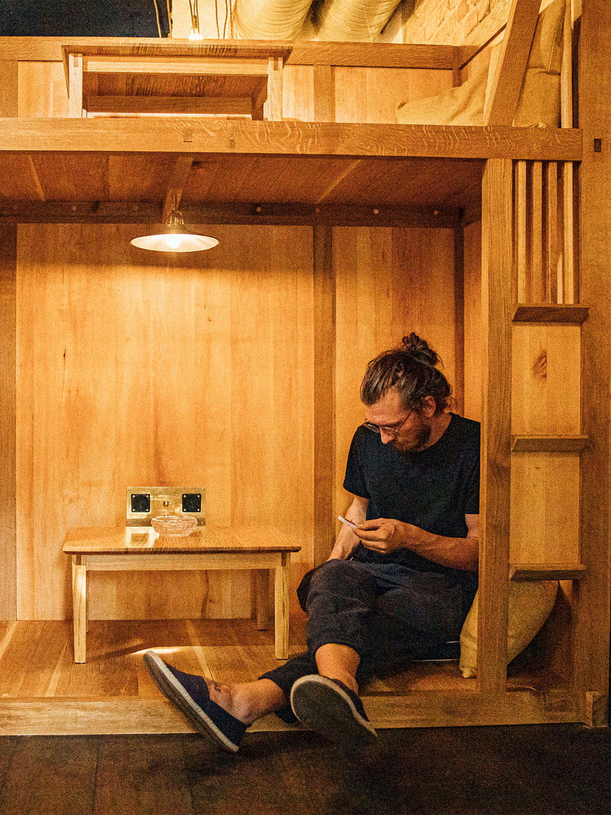 Eine Person sitzt in einer atmospährischen hölzernen Nische und schaut auf ein Mobiltelefon. Die Nische ist mit einem Tisch, einer Lampe und Steckdosen ausgestattet. Gebaut wurde die Sitznische des Multitude Hostels von Mimikry, designbüro in Leipzig