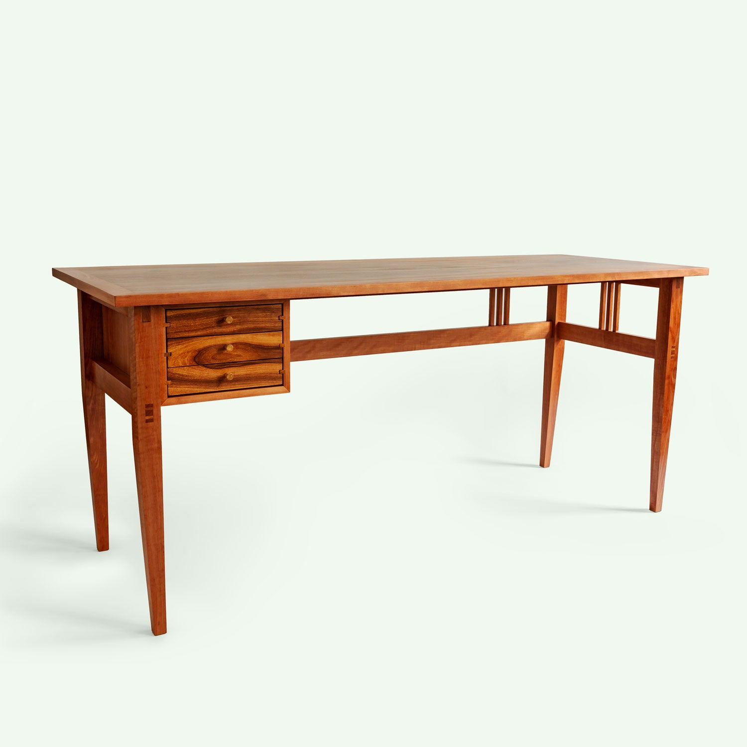 Der Schreibtisch wird aus einem schrägen Winkel gezeigt, der die gesamte Form und Struktur des Tisches betont. Die Kombination aus Apfelholz und Elsbeere verleiht dem Tisch ein einzigartiges und edles Aussehen.