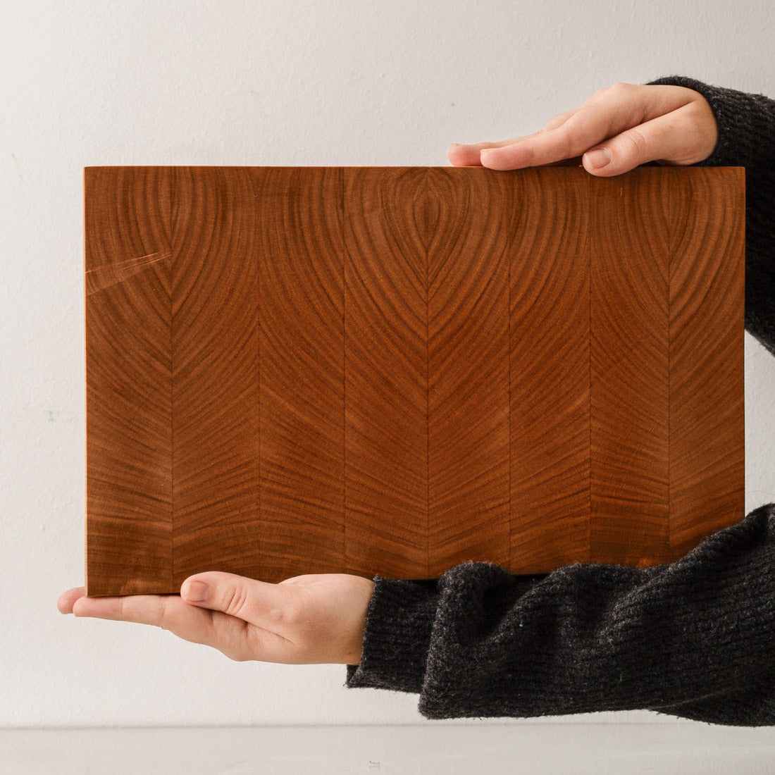 Großes Hirnholz Schneidebrett Kirsche in einer frontalen Lifestyle-Aufnahme, horizontal von zwei Händen in einem dunklen Strickpullover präsentiert, was die beeindruckenden Ausmaße, das symmetrisch gespiegelte, wellenförmige Muster der Jahresringe im warmen Stirnholz und die edle Oberflächenqualität des Massivholzes zeigt. Handgefertigt von der Tischlerei Mimikry in Leipzig.