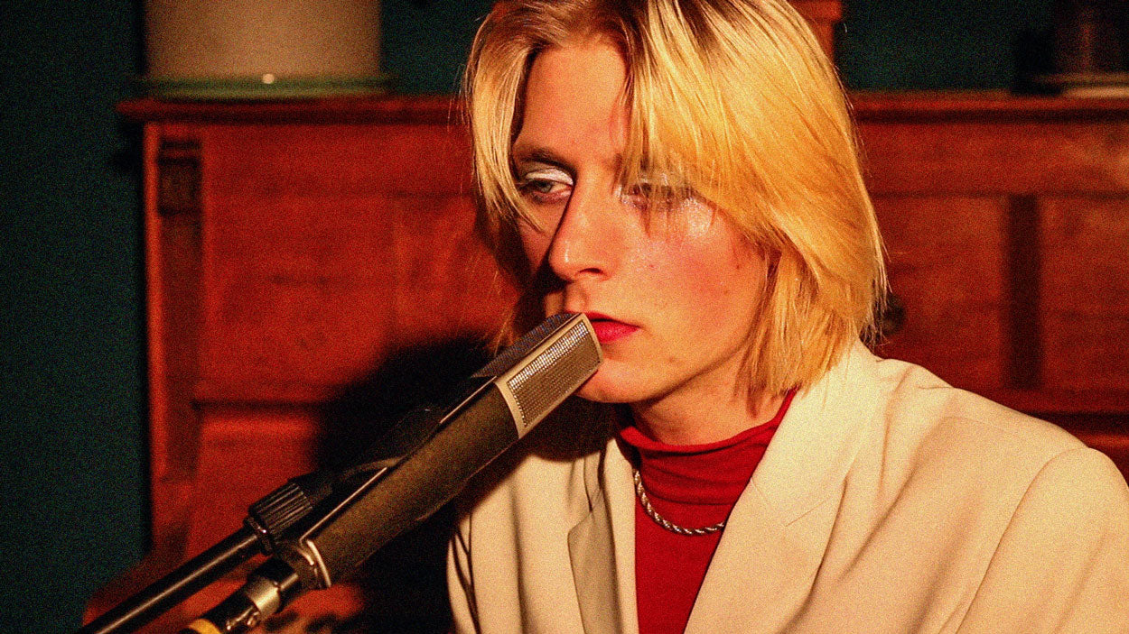 Kat Embo zeigt eine stimmungsvolle Live-Session-Aufnahme im Innenraum der Tischlerei mimikry in Leipzig, fokussiert auf ein Porträt des Künstlers mit blonden Haaren, markantem Augen-Make-up, hellem Sakko und rotem Rollkragenpullover direkt am Mikrofon, um die emotionale und fokussierte Performance vor einem hölzernen Klavier im Hintergrund zu veranschaulichen.
