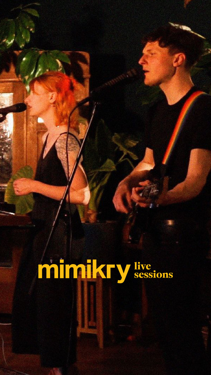 Twins in Colour zeigt eine stimmungsvolle Live-Session-Aufnahme im Innenraum der Tischlerei mimikry in Leipzig, fokussiert auf das Duo mit einer rothaarigen Sängerin im Profil und dem Gitarristen mit regenbogenfarbenem Gitarrengurt an den Mikrofonen, um die lebendige und intime Konzertatmosphäre vor Zimmerpflanzen im Hintergrund zu veranschaulichen.02