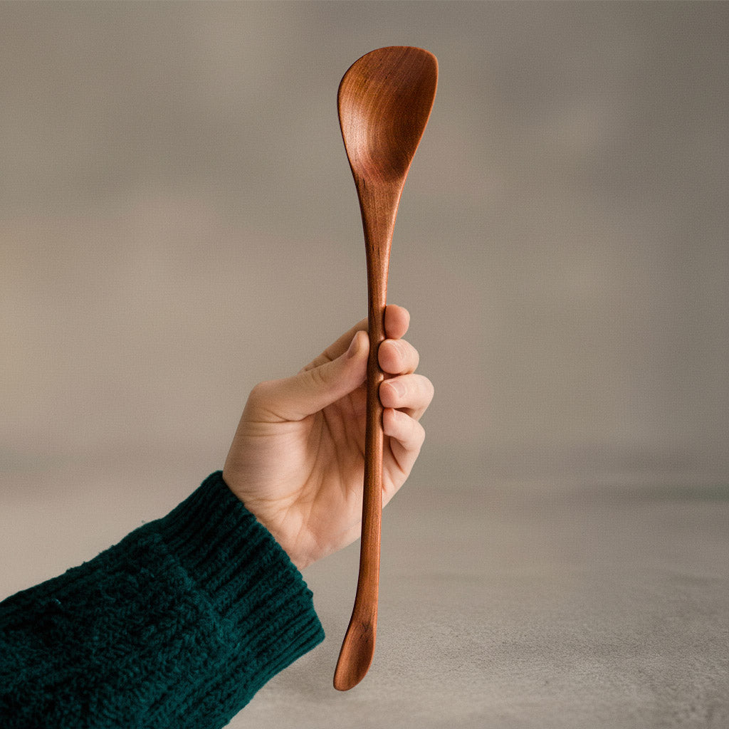 Hand hält handgefertigten asymmetrischen langen Löffel aus Elsbeere mit elegant geschwungenem Design und glatter Oberfläche, nachhaltig produziert, perfekt zum füttern von Babys