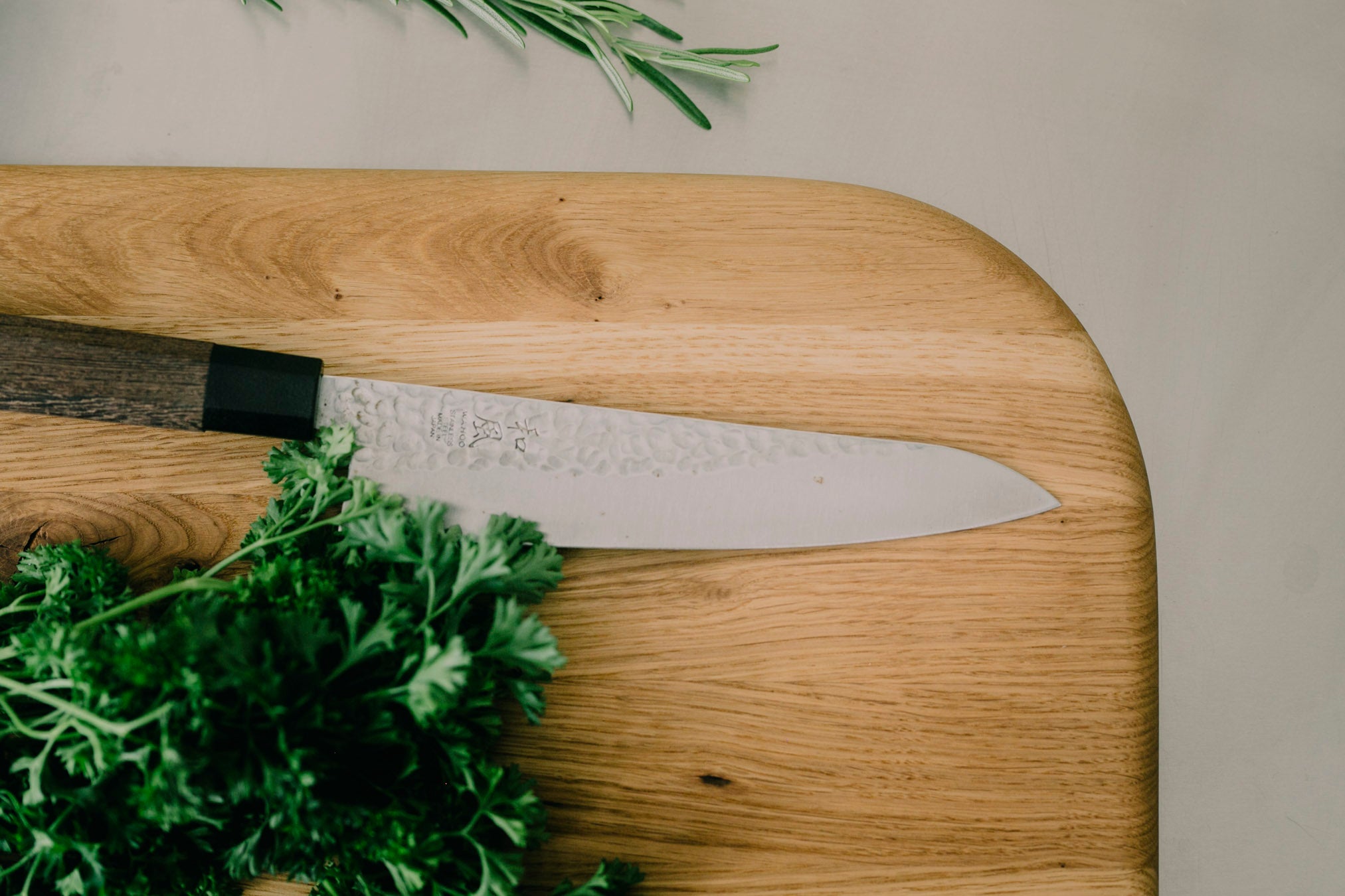 Draufsicht auf die verrundete Kante eines Holz Schneidebrettes aus Eiche, auf dem ein japanisches Messer und frische Petersilie liegt.